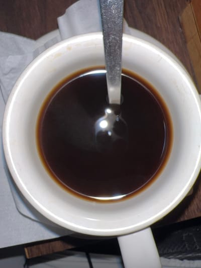 The image displays a white ceramic mug, viewed from an overhead perspective, filled with a dark brown liquid, consistent with coffee. A silver-toned metal spoon is positioned vertically within the liquid, with a distinct circular light reflection visible on the liquid's surface. The inner rim of the mug shows minor brown discoloration. The mug is resting on a surface that includes a crumpled white paper napkin to the left and a dark brown wooden surface, likely a table or countertop, to the right. No individuals are present. The environment is an indoor setting. This scene is situated in Toyapakeh, Indonesia.