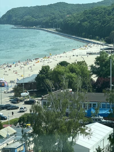 This is an aerial, eye-level, daytime, wide shot of the city of Gdynia, Poland, specifically the beach area, looking southeast. The image captures a bustling beach with numerous people sunbathing, swimming in the sea, and walking along the shore. A paved promenade runs along the edge of the beach, with trees lining the inland side. Several buildings, including what appear to be beachside facilities and possibly some sort of resort or restaurant, are visible behind the promenade. Further inland, more trees and a hilly landscape form the backdrop. In the foreground, there is a parking lot with several cars and some tents, suggesting the presence of a commercial or event area. A body of water, likely the Baltic Sea, occupies the upper left portion of the image.