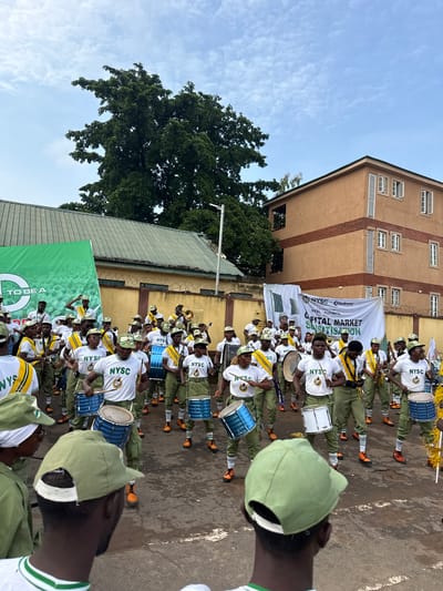 In Alagba, Nigeria, a group of young people in matching green and white uniforms, some wearing hats, participate in a parade. The individuals are playing various musical instruments, including drums and brass instruments, and appear to be part of a marching band. They are standing on a paved surface, possibly a street or courtyard, with a large building and a green banner in the background. The banner displays text in English, including "NYSC" and "CAPITAL MARKET SERIALISATION PROGRAM." The sky is cloudy.