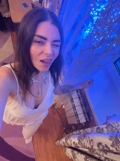 The image, captured indoors, displays a young woman in what appears to be a bedroom or living space in Krasnoe-na-Volge, Russia. The woman, with dark hair, is wearing a white slip with lace detailing and white pajama pants, along with white socks. She is winking at the camera and sticking her tongue out, suggesting a playful or casual moment. She is adorned with a multi-layered necklace featuring various charms.

The environment is characterized by warm wooden flooring and a patterned rug in shades of pink and purple. A radiator is visible in the background, along with a patterned curtain that is illuminated by blue string lights, creating a vibrant and atmospheric lighting effect. The overall impression is of a personal, intimate setting within a home.