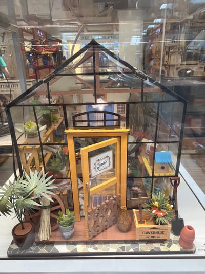 This is a top-down, eye-level, medium shot of a miniature greenhouse display, viewed through glass, possibly in a museum or store. The greenhouse itself is made of clear acrylic panels with black framing, and it is filled with various miniature gardening-related items.
Inside the greenhouse, there are shelves lined with tiny potted plants, some with vibrant green leaves and others with delicate white and yellow flowers. A miniature wooden crate labeled "FLOWER HOUSE" sits on a shelf, holding a bouquet of artificial orange and yellow flowers. A small toy shovel rests on top of the greenhouse, and a tiny birdhouse with a blue decorative element is also visible. Other details include a small wooden wagon wheel, bundles of straw, and various decorative elements that mimic gardening tools and accessories.
In the background, reflections show what appears to be a larger display or exhibit, with more complex structures and objects, possibly representing a city or industrial scene. The lighting is bright and appears to be artificial, likely from indoor display lighting. The overall scene evokes a sense of intricate craftsmanship and a miniature world dedicated to gardening. There is no visible text on the greenhouse itself, but the "FLOWER HOUSE" label on the crate is legible. The reflections in the glass obscure some details of the background display.