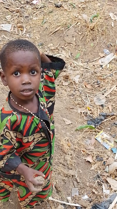 A young child with dark skin and short hair stands in a dirt field littered with debris. The child wears a colorful patterned shirt and a beaded necklace. The child’s eyes are wide and expressive. One hand is raised to the head while the other holds a clump of dirt. The background shows dry grass, leaves, and scattered trash. The overall impression is one of poverty and resilience.
