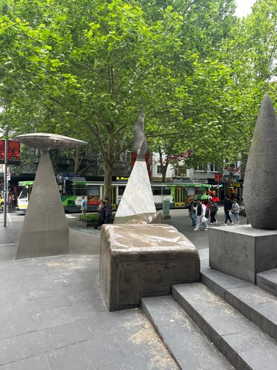 An outdoor scene shows an urban plaza with geometric concrete structures. Trees with green leaves frame the left side. Several people are standing near a green and white tram that is on the road in the background. Graffiti marks one of the concrete structures. The image is shot from a low angle.