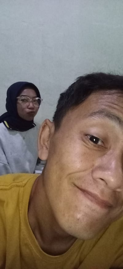 Two individuals are captured in a close-up, selfie-style photograph. The person in the foreground, on the right, is a male whose face fills the majority of the frame, with a yellow t-shirt visible on his chest. His right ear is also prominently displayed. Behind him, to the left, a female wearing a black hijab and glasses is visible. She is looking towards the camera with her lips pursed. The background consists of a plain, light-colored wall. The lighting appears to be artificial and consistent. The setting is indoors, likely a residential space.