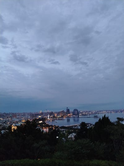 This is an outdoor, high-angle, medium shot captured at dusk. The scene is a cityscape overlooking a body of water, likely a bay or sea, with a skyline dotted with various buildings. In the foreground, dark green trees and foliage dominate the lower portion of the frame, creating a natural barrier between the viewer and the urban landscape.

The city lights are beginning to illuminate the buildings, creating a warm contrast against the cool, muted tones of the sky. The sky itself is overcast with a pattern of dark and light gray clouds, suggesting a cloudy evening. The water reflects the lights from the city, creating a shimmering effect. In the mid-ground, several prominent structures can be seen, including what appears to be modern architecture with distinctive shapes. The overall mood is serene and perhaps a bit dramatic due to the twilight setting and the presence of the clouds.

The location cues, such as the architecture and the context provided, point to Sabail Raion, Azerbaijan. There is no visible text in the image. The scene captures the transition from daylight to night in an urban coastal environment.