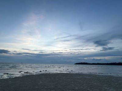 This is a serene, wide-angle shot of a tranquil beach and ocean scene, likely taken at dusk or dawn given the soft, diffused light and the predominantly blue and grey hues. The sky is a gradient of pale blue at the top, transitioning to lighter, overcast tones with wispy clouds closer to the horizon.
The foreground is dominated by damp, rippled sand, indicating a receding tide. Scattered dark shapes, possibly seaweed or debris, are visible on the wet sand where the water meets the shore. The middle ground is the calm ocean, with gentle, horizontal waves creating a striped effect. In the distance, a faint, dark line of land with some vegetation and structures can be seen, suggesting a coastline or an island. Another, smaller landmass or island appears on the far left horizon.
The overall mood is peaceful and contemplative, evoking a sense of solitude and natural beauty. The lighting suggests either early morning or late evening, with the sun not directly visible. There is no visible text or people in the image. The context provided places this scene in Ko Pha-ngan District, Thailand, a location known for its beaches and tropical environment.