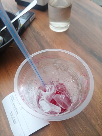 A close-up, overhead shot captures a clear plastic cup filled with ice and a red liquid, possibly a beverage. A blue and white striped straw extends from the cup. The cup sits on a wooden table, and condensation has formed on its exterior. To the left of the cup, a small portion of a black bowl containing food and a silver spoon is visible. Behind the cup, a glass of water and a pack of cigarettes are also on the table. In the foreground, a white receipt is partially visible beneath the cup. The receipt shows the text "Pesanani 2 Indomie Goreng Original" followed by a date and time, "19 Jun 26 17:05," and "dicetak: AMAR." The scene appears to be set in Geulumpangsulu Timu, Indonesia.