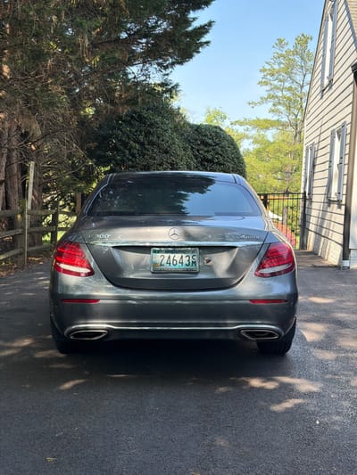 A grey Mercedes-Benz E300 sedan is prominently featured, parked on a paved surface with its rear facing the viewer. The car displays a Mercedes-Benz logo centrally on the trunk, with "E 300" badging on the lower left and "4MATIC" on the lower right. It has dual, rectangular exhaust outlets integrated into the rear bumper.

A Maryland license plate is visible, showing the alphanumeric code "24643M". The plate also reads "MARYLAND '26" at the top and "JOHNS HOPKINS ENGINEERING" at the bottom, indicating a specialized plate.

The setting is outdoors in what appears to be a residential area during a bright, sunny day. A clear blue sky is visible in the upper left quadrant. Sunlight casts dappled shadows of overhead trees onto the dark asphalt surface to the right of the car. In the background, a light-colored house with horizontal siding and a dark metal fence are present. Abundant green foliage, including tall trees and dense bushes, frames the scene, with some bare tree branches visible towards the lower left. A low wooden barrier or fence is also partially seen on the left. The scene depicts a stationary car in a driveway or parking area.