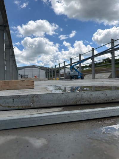 A construction site in Janowice, Poland, displays the initial stages of a building's frame. The foreground is dominated by a wet concrete surface, with two horizontal metal beams resting on it. The beams show signs of wear, with some rust and dirt accumulation. To the left, a rectangular piece of weathered wood lies on the concrete.
In the mid-ground, the skeletal structure of a building is taking shape, with vertical steel columns supporting horizontal beams. A blue boom lift, a piece of heavy machinery used for elevated access, is parked near the structure. The area surrounding the construction is partially covered with concrete, with some sections appearing wet and reflecting the sky.
The background features a large, partially completed building with a white, corrugated metal exterior. Further in the distance, a tree-lined slope rises, and the sky is filled with a mix of white, puffy clouds against a bright blue expanse. Two individuals, one wearing an orange vest, are visible near the partially completed building in the distance, suggesting ongoing construction activity. The overall scene depicts an active construction zone under a bright, cloudy sky.