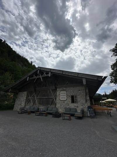 The image shows an exterior view of the "SALZSTADEL RESTAURANT" in Reutte, Austria. The building features a stone facade and a large wooden roof structure with visible timber framing. In front of the building, there are several outdoor seating areas with dark gray sofas and wooden tables. A chalk A-frame sign stands near the entrance. To the right, there are more outdoor seating areas with wooden benches and tables under white umbrellas. A forested hillside rises behind the building. The sky is overcast with dramatic, dark clouds.