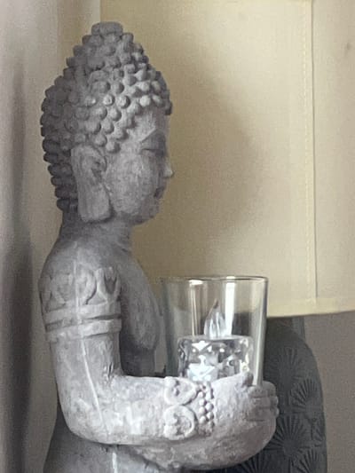A close-up, side-view shot shows a grey, textured statue of a Buddha figure. The Buddha is holding a clear glass votive holder containing a clear, faceted candle with a visible flame. The candle appears to be electric. The statue's arm and hand are visible, adorned with beaded bracelets and carved bands. The background consists of a plain, cream-colored surface with a visible seam, possibly a lampshade, and a darker grey textured object on the right. The image is an indoor still life, with no discernible location in Bunbeg, Ireland.