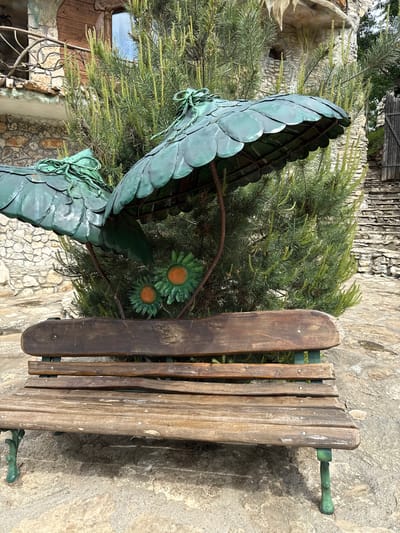 A medium shot shows an outdoor scene featuring a wooden park bench adjacent to a sculpted metal plant. The bench, made of aged wood planks attached to green metal supports, sits on a stone surface that extends into the background. Next to the bench stands a metal sculpture of plant leaves with flowers in green and orange hues. Between the bench and metal sculpture is a small pine tree. The background includes stone walls and parts of buildings, indicative of an urban or park setting in Lukovit, Bulgaria.