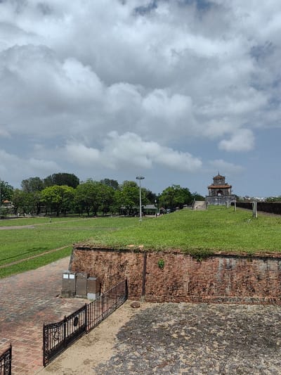 The image depicts an expansive outdoor scene within the Imperial City of Hue, Phú Xuân Ward, Vietnam. The sky occupies the upper two-fifths, featuring dense light gray and white cumulus clouds with patches of clear blue sky.

In the foreground, a dark, rough ground surface covers the lower right. To the left, an ornate black metal fence extends, bordering a reddish-brown brick pathway. Three metallic utility boxes are positioned against a weathered brick wall that runs horizontally across the mid-ground. This brick wall shows signs of discoloration and minor vegetation growth.

Above the brick wall, a large, well-maintained green grassy lawn extends into the background, sloping gently upwards towards the right. A reddish-brown brick path is visible on the far left of the grassy area. A line of dense green trees, interspersed with smaller structures and a tall, multi-headed streetlamp, stretches across the mid-distance. Several small human figures are discernible walking on the grass and near these distant structures.

Prominently situated on an elevated grassy mound on the right side of the image is a traditional Vietnamese multi-tiered pagoda-like structure. It features dark grey walls, a distinctive reddish-orange tiled roof with curved eaves, and a central arched opening. A set of steps leads up to this structure. Faint outlines of distant urban buildings are visible on the far right horizon.