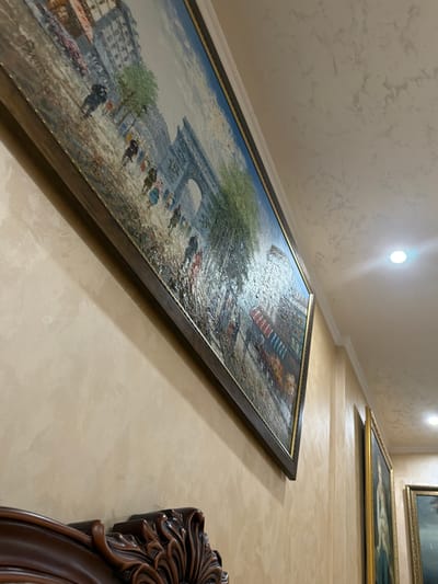 A large oil painting of a Parisian street scene hangs on a textured, beige wall. The painting, framed in dark wood with a gold inner trim, depicts people walking past buildings and an archway under a cloudy sky. The artwork is angled, showing its side and a portion of the wall, which has subtle, darker textured patterns. To the right, a second framed painting with a gold frame is visible, and further to the right, a third painting with a similar gold frame is partially visible. The ceiling, also beige and textured, features a white molding and a bright recessed light fixture. In the lower left corner, the ornate, dark wood carving of a piece of furniture is visible. There is no identifiable information in the image to suggest a location in Vesyoloye, Russia.