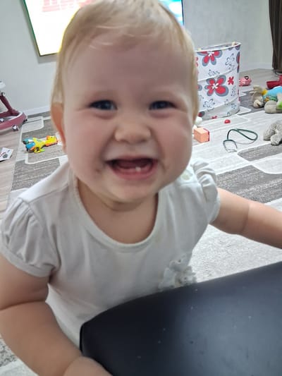 A close-up, head-and-shoulders shot of a smiling baby is captured from a slightly low angle. The baby, with fair hair and wide, dark eyes, is wearing a white t-shirt with puffed sleeves. The background shows a carpeted floor with a gray and white geometric pattern. Scattered on the carpet are various toys, including a yellow and blue toy airplane, a small pink toy pig, a plush gray stuffed animal, and a toy stethoscope. A cylindrical toy bin with a floral pattern of red and white flowers is also visible. To the left, a portion of a red and silver baby walker is visible, and a television screen showing pink and white text is partially seen in the upper left corner. A dark, flat surface, possibly a table or bench, occupies the lower right portion of the frame. The scene appears to be an indoor play area within a residential setting. The image does not provide specific information to determine its location within the city of Semey, Kazakhstan.