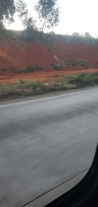 The image displays an exterior roadside view, captured from within a moving vehicle in Kikuyu Township ward, Kenya. A dark, curved vehicle interior component partially frames the bottom right. A grey asphalt road, exhibiting horizontal motion blur, extends across the lower-mid portion, featuring a continuous white lane marker on its right edge. To the immediate left of the road is a narrow dirt verge interspersed with sparse green vegetation and scattered light-colored debris. Directly behind this verge, a prominent, steep reddish-brown earthen embankment rises, partially covered with green foliage and scrub. Deciduous trees are silhouetted against a bright, hazy sky at the crest of the embankment. No individuals, animals, or other vehicles are visible.