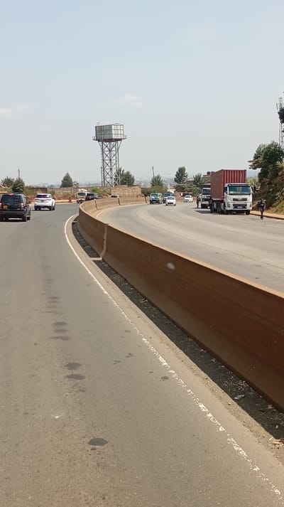 The image displays a multi-lane asphalt highway in Gitaru ward, Kenya, under a hazy, light blue sky. The road curves gradually to the left from the foreground perspective. Two traffic lanes are visible moving away from the camera. On the left lane, a dark SUV, followed by a white SUV, and then a white minibus, are in motion. On the right side of the road, a white semi-truck with a red shipping container is positioned on the shoulder or outer lane, followed by a second white truck. Two individuals are present on the right; one person in dark clothing walks on the far right shoulder, and another stands near the cab of the container truck. A curved, brown concrete barrier runs along the right side of the immediate roadway, bordering a dirt strip. In the mid-ground, a tall metal water tower with a rectangular tank is visible on the left, positioned above a vegetated embankment and distant urban structures. Scattered trees and bushes line the road and background. The asphalt surface exhibits several dark, irregular stains.
