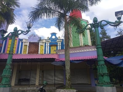 The image displays an outdoor urban scene in Gadog, Indonesia, featuring a row of multi-story buildings and prominent street infrastructure under a partly cloudy sky. In the foreground, two ornate, dark green streetlights with decorative scrollwork and double lanterns frame the view. A black motorcycle with a green helmet resting on its seat is parked in the lower center. The buildings behind feature a uniformly pink ground floor with closed metal roller shutters over storefronts. A small, dark table is visible in front of one shuttered shop. The second story comprises varied, brightly colored facades: yellow with blue vertical stripes on the far left, a purple section with a stepped gable, and a blue section with a similar stepped gable and white vertical trim. A distinct green tower-like structure with white vertical fluting and a red domed roof stands prominently on the right side of the building complex, partially obscured by a central palm tree. Additional palm fronds extend into the upper left, and other trees are visible in the background to the right, beneath a dark, framed canopy structure. No individuals are present, and no actions or interactions are depicted within the static environment.
