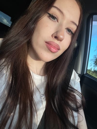 This is a close-up, selfie-style image of a young woman inside a vehicle, likely a car, as suggested by the seatbelt and interior. The woman has long, dark brown hair that is flowing loosely. She is wearing a white t-shirt and a silver pendant necklace with a heart shape. Her makeup includes winged eyeliner and lip gloss. The lighting suggests it is daytime, with sunlight casting shadows and highlights on her face and hair. The background shows a glimpse of the sky and trees through the car window, indicating an outdoor setting. The overall mood is relaxed and casual. There is no visible text in the image.