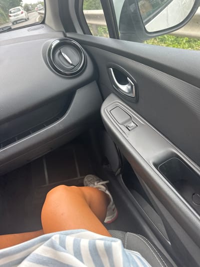 This media file captures a close-up, high-angle view from inside the passenger side of a car, focusing on the door panel, footwell, and a seated passenger. The scene is brightly lit by natural daylight, suggesting it is daytime.
A person's right arm, bent at the elbow, and their lower leg with a foot wearing a light gray and red sneaker are visible in the lower right and bottom center of the frame. The person is wearing a light-colored garment with thin blue and white vertical stripes.
Key objects within the car interior include a dark gray, textured plastic door panel. Prominently featured on the door are a chrome-finished interior door handle, an electric window control switch with a small window icon, and a circular air vent with a chrome ring. A side mirror reflects a view of a road or street, green foliage, and other vehicles in the distance, indicating the car is outdoors, possibly in motion or parked alongside a road. Part of a dark car seat with white stitching is visible on the right. Storage compartments and cubbies are visible along the door and below the dashboard.
No specific activity beyond being seated as a passenger in a vehicle is depicted. The overall impression is that of a journey in Podličak, Montenegro, under clear weather conditions.