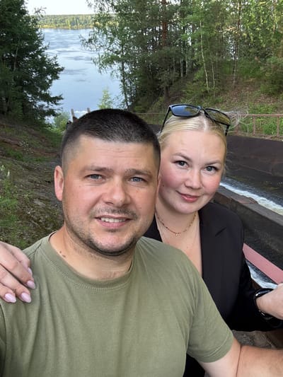 This media file is a selfie featuring a couple. The man, in the foreground, is wearing a green t-shirt and has short, dark hair and stubble. He is smiling at the camera. The woman is behind him, wearing a black top and a necklace. She has blonde hair and is also smiling. She is wearing sunglasses on top of her head. The background shows a body of water with trees and greenery on its banks, suggesting a natural setting. There is also a concrete structure with water flowing over it, possibly a small dam or spillway. The scene appears to be outdoors during the daytime, with natural lighting. The overall mood seems relaxed and happy, indicative of a casual outing or vacation. The image was taken in Ol'khovskoye, Russia. No text is visible in the media file.