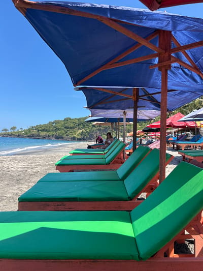 This is a photograph taken at a beachside resort in the city of Karangasem, Indonesia. The image shows a row of empty green lounge chairs with orange frames lined up on a sandy beach, positioned under large blue umbrellas. In the background, the ocean meets the shore with gentle waves, and a lush green hillside dotted with trees rises from the coastline. The sky is a clear, bright blue. A few people are visible in the distance, seated on lounge chairs under other umbrellas, and some are in the water. The perspective is low, focusing on the foreground lounge chairs and umbrellas, with the beach and ocean stretching out to the left.