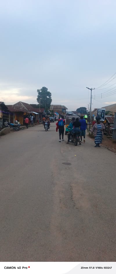 The image depicts a street scene in Barikin Gidar Kanat, Nigeria, likely in the late afternoon or early evening, given the soft, diffused light and overcast sky. The scene is bustling with activity, showcasing a typical day in a Nigerian town.
The main subjects are the people navigating the street. Several individuals are on foot, including a person in the foreground wearing a blue and red backpack and another person further down the road in a patterned dress. Others are riding motorcycles or a three-wheeled auto-rickshaw, suggesting various modes of transportation are in use.
Objects in the scene include parked motorcycles and the auto-rickshaw. The buildings lining the street appear to be small shops or homes, with corrugated iron roofs, some of which show signs of age. A large tree stands prominently in the background, and utility poles with electrical wires are visible. A billboard featuring a political candidate or advertisement is also present on the right side of the street, with text that is difficult to fully decipher but appears to say "SAMUEL DIGITAL."
The overall atmosphere is one of everyday life and commerce. The people's expressions are not clearly discernible, but the activity suggests a normal, active day. The road is paved and shows signs of wear. The weather appears to be cloudy or hazy, contributing to the subdued lighting.
Visible text includes "CAMON 40 Pro" and "23mm f/1.8 1/100s ISO247" at the bottom of the image, which are likely camera specifications. The text on the billboard is partially visible.