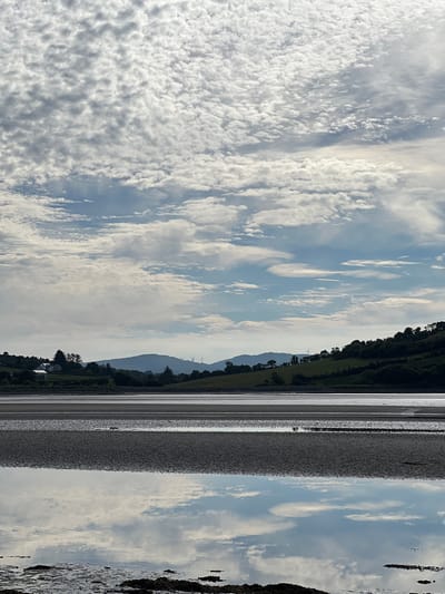 The image depicts a scenic view of a body of water in Dunuisce, Ireland. The foreground is dominated by calm water reflecting the sky, with patches of exposed mudflats or sandbanks in horizontal bands. The water's surface is smooth, creating a mirror-like effect for the sky and distant landscape.
In the middle ground, a sandy or muddy shoreline stretches across the frame, featuring distinct textural variations. Beyond this, a gently sloping hillside is covered with lush green vegetation, dotted with trees and a few white buildings. In the far distance, rolling hills form a silhouette against the sky, with several wind turbines visible on the horizon, indicating the presence of renewable energy infrastructure.
The sky above is filled with a dramatic display of clouds. There are numerous small, puffy white clouds scattered across a light blue sky, interspersed with larger, more diffused cloud formations and streaks of cirrus clouds. The lighting suggests it is daytime, likely late morning or early afternoon, with the sun casting a soft glow on the landscape. The overall atmosphere is serene and natural, capturing a tranquil moment in the Irish countryside.