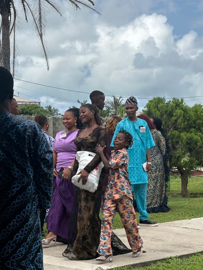 In Itoga, Nigeria, a group of people are gathered outdoors on a cloudy day, possibly for a celebration or event. A woman in a purple dress and a woman in a dark lace dress are talking and smiling. The woman in the lace dress is holding a white bag with a logo that reads "Elegance Hospitals". To their right, a man in a bright blue traditional outfit, wearing a patterned headwrap, stands next to a young child in a colorful patterned jumpsuit. The child is laughing and looking up. In the foreground, a person in a dark blue and black patterned garment is partially visible. Behind the group, a palm tree and lush green vegetation are present. A fence and part of a building are visible in the background, suggesting a residential or community setting.