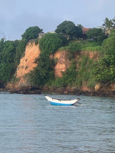 A wide-angle, eye-level shot captures a serene scene in Mirissa, Sri Lanka. In the foreground, the blue and white hull of a small fishing boat floats gently on the calm, gray-blue water. The boat appears to be at rest, with two wooden outriggers extending from its sides. Beyond the water, a steep, reddish-brown cliff rises, covered in lush green vegetation. The cliff face shows signs of erosion, with layers of soil exposed. Atop the cliff, several trees and a building with a brown-tiled roof are visible. The sky is overcast, creating a diffused light that softens the entire scene. The image conveys a sense of tranquility and the natural beauty of the coastal landscape.
