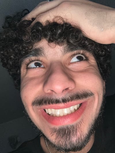 An extreme close-up, low-angle portrait depicts a male individual, appearing to be in his 20s or 30s, with a broad, open-mouthed smile revealing both upper and lower teeth. He possesses dark, tightly curled hair, a mustache, and a goatee-style beard. His dark eyes are directed upwards and to the right, with visible long eyelashes and eyebrows. The individual's right hand is positioned at the top of his head, with fingers grasping his hair. He is wearing a dark-colored garment and a thin, silver-colored chain or necklace is visible below his chin. The background is uniformly dark and lacks distinct features, suggesting an indoor setting with minimal ambient light. This scene is located in Marcoussis, France.