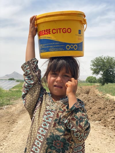 A young girl is depicted outdoors, carrying a large yellow bucket on her head under a bright, clear sky. She is positioned slightly to the right of the frame's center, looking towards the viewer with a neutral or slightly strained expression. Her left hand is raised, steadying the bucket, while her right hand is near her ear. She wears a patterned long-sleeved dress in shades of beige, brown, green, and light blue, and several layered necklaces, including a clear, multi-faceted one and a turquoise beaded one.
The yellow bucket prominently displays the text "GREASE CITGO" and "CALCIUM BASED GRADE 3". Smaller text on the bucket, though partially obscured, describes the properties and uses of the grease, mentioning "thickener preferable structure," "protect metal surfaces in wet operating conditions," "Chassis Grease Point Lubrication," and "Industrial Rolling Element Bearings."
The background suggests a rural or semi-rural setting. To the girl's left, a dirt path or road leads past green fields, with multiple solar panels visible in the distance, indicating some infrastructure. Beyond the solar panels, a mountain range is faintly visible under the hazy sky. To her right, a large mound of excavated earth or dirt is piled up, contrasting with the greener vegetation further back and a few scattered trees. The sunlight suggests it is daytime. The scene is set in Ashoqah, Afghanistan, depicting an everyday activity in this environment.