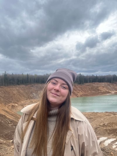 A photograph depicts a woman in Ivlevo, Russia, standing by a quarry or a water-filled excavation site. The woman is wearing a beanie and a light-colored coat. The background shows an industrial landscape with a water body and exposed earth. The sky is overcast.