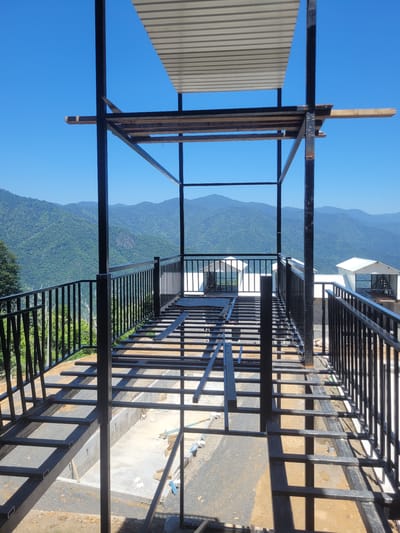 The image shows an unfinished construction site with a view of a mountainous landscape. A metal structure, likely part of a balcony or walkway, dominates the foreground. Corrugated metal sheeting is visible on the left side, partially covering the structure. The background reveals lush green mountains under a clear blue sky. The sun appears to be high in the sky, casting distinct shadows from the metal beams and railings. The ground below the structure is a mix of dirt and gravel, with some evidence of construction materials such as concrete and what appear to be discarded cardboard boxes. The scene suggests an outdoor setting in a rural or semi-rural area, possibly a residential or resort development, given the scenic mountain backdrop. There are no people visible in the image. The overall impression is one of ongoing construction against a natural, serene backdrop. The rotation of the image makes it difficult to ascertain specific details about the construction phase, but it is clearly in progress.
