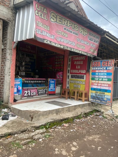 The exterior of a small storefront, located at latitude -7.021 and longitude 107.675, in the city of Baleendah, Indonesia, is visible. The shop's name is "Lita Cell," and it appears to be a cell phone repair and accessory shop. The storefront is decorated with colorful signage advertising the various brands and services offered, including "Serpis HP," "Lita Cell Service HP," and "Xiaomi, Asus, LG, Coolpad, Hardware, Motorola, Mito."  The store is decorated with a red, white, and yellow color scheme, with various brands of cell phone cases and accessories visible in the window display. Two plastic chairs sit in front of the store, indicating the presence of people waiting to be served. The shop front is set back from the sidewalk, with an open concrete area in front, which is unkempt, and littered with trash.  There is no one currently in the immediate area of the store.  There is a plastic bag of trash on the ground next to the storefront, and various pieces of litter are scattered on the sidewalk. There is green moss growing in the cracks of the concrete sidewalk. The weather appears to be overcast.  The storefront is built in a similar style to the other buildings in the area, with a concrete facade and a simple design.  There is a visible power line running across the top of the image, which appears to be above the storefront and behind the signage. The storefront appears to be a small, local business that provides services and products for the surrounding community.  The scene is a typical example of a small, local shop in Indonesia.