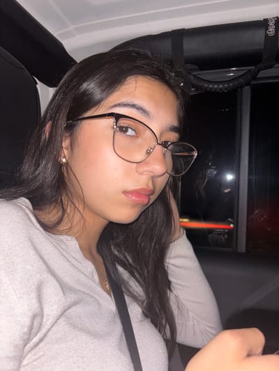 A female is lying in the back of a Jeep vehicle. She wears eyeglasses, has dark brown hair, and a fair complexion. She is wearing a white-colored top. The Jeep is implied by the visible "Jeep" branding on the grab handle. The environment suggests it is nighttime, and blurred lights are visible through the vehicle's window. Location: San Antonio, United States of America is unverifiable by the image itself, but it was requested that it is included.