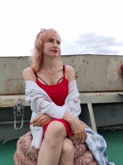 A woman with peachy-blonde hair and bright red lipstick is seated outdoors. She is wearing a red spaghetti-strap dress and a white cardigan with a patterned design. A silver necklace with a small pendant is visible around her neck. Her arms are crossed, and she is wearing a ring on her right hand. She is sitting on a large, textured, light pink object, possibly a cushion or a large rope. To her right, a blue and white patterned scarf is draped. Behind her is a rusty, metallic surface, suggesting an industrial or outdoor setting. The background shows a cloudy sky. The image appears to have been taken during the daytime. The location is described as Bereg, Russia, though the specific landmarks within the city are not discernible from the image itself.