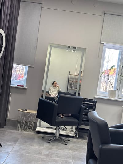 This image depicts the interior of a beauty salon or similar establishment in Gamovo, Russia. A large, illuminated, floor-standing mirror is the central object, reflecting a female individual. This individual, wearing a light-colored long-sleeved top and light trousers, is seated on a dark bench or sofa behind a black salon chair. She holds a white smartphone in her right hand, oriented towards the mirror, indicative of a selfie or self-observation. Her dark hair is pulled back.
In the foreground, a black hydraulic salon chair with a chrome base faces away from the viewer, with a patterned fabric or bag resting on its seat. A second black salon chair is partially visible on the right. The room features light grey walls and grey square-tiled flooring. Two windows with light grey roller blinds are present; one on the left is partially covered by dark grey curtains, and the other on the right displays a potted plant on its sill. Through the windows, bare trees and distant structures with red-orange elements are visible under ambient daylight. A small, round, white wireframe side table near the left window holds a dark object, likely a phone. A white ring light on a stand is partially visible on the far left. In the reflection, a dark-framed shelving unit with multiple compartments holds various items behind the seated individual. Two crescent-shaped indentations are visible on the ceiling above the mirror.