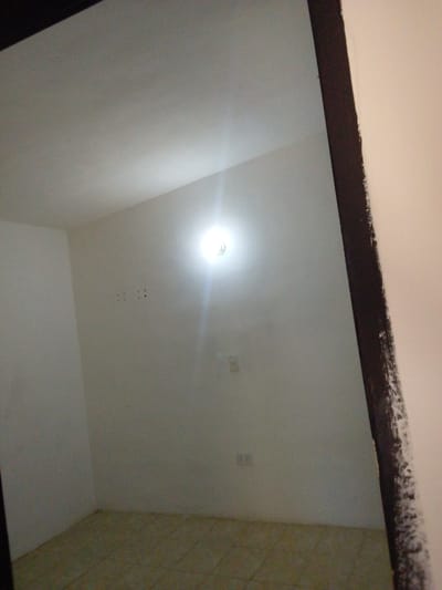 This is a low-angle, indoor photograph of an empty room with white walls and a tiled floor. A single, bright light bulb hangs from the ceiling, illuminating the space. To the left, a dark, rough-textured doorway or frame bisects the image diagonally, leading into what appears to be another room or hallway.

On the far wall, there are two electrical outlets, one above the other, and a cluster of four small, dark marks that could be holes or remnants of a fixture. The overall impression is of a simple, undecorated living space, possibly recently vacated or awaiting furnishings. The lighting is harsh, with a glare from the bulb dominating the central part of the image. There is no visible text in the image. The location context provided is El Maco, Venezuela.