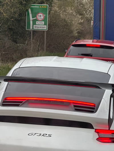 The image presents a forensic view of vehicular traffic on a road near Stratford-on-Avon, United Kingdom. In the immediate foreground, the rear of a white Porsche GT3 RS is visible, featuring a prominent carbon fiber rear wing. The vehicle's full-width LED taillight bar is illuminated red, displaying the "PORSCHE" emblem, and the "GT3RS" model designation is visible on the lower rear fascia. Moisture droplets are present on the car's upper surfaces.

Immediately ahead of the Porsche, partially obscured, is the rear of a red SUV or crossover vehicle, also with illuminated rear lights and a visible rear wiper blade. The vehicles appear stationary or in slow-moving traffic.

In the background, to the left, a green road sign on dual posts provides directional information: "Coventry A423" is indicated with a left-turn arrow, under which a red circular sign displays an "18t mgw" (Maximum Gross Weight) restriction. Below this, "2/3 mile" and "Stockton" are listed, associated with the left turn. The surrounding environment consists of bare or sparsely foliated trees and shrubs lining the roadside under an overcast sky.

Based on the road sign information ("Coventry A423," "Stockton," and the general direction implied by the A423), the scene is located on the A422 (Banbury Road) as it approaches the junction with the A423 and B4086, specifically near Hatton Rock, approximately 6-7 miles east of Stratford-upon-Avon town centre, within the Stratford-on-Avon district.