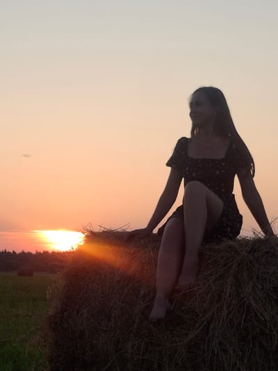 A silhouette of a young woman in a dark floral dress sits on a large hay bale in a field at sunset. The sky is a gradient of pale yellow and pink. The sun is low on the horizon, casting a warm glow over the scene. The woman is looking to her left, with her legs crossed. The hay bale is rough and textured, with strands of hay visible. In the background, there is a dark line of trees and another hay bale in the distance. The overall impression is one of peace and tranquility. The location is the outskirts of Perevoznoe, Russia.
