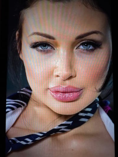 The scene is located in the city of Balykchi, Uzbekistan. The image is a close-up, head-and-shoulders portrait of a female individual facing directly forward. She has fair skin, blue eyes, dark eyebrows, and visible dark hair. Her eyes are accentuated with dark eyeliner and long, artificial eyelashes. Her full lips are coated in a glossy pink lipstick. A dark fabric with a white striped pattern, resembling a scarf or a garment's collar, is draped around her neck and upper chest area. The background is uniformly dark and out of focus, providing no specific environmental details. The individual is stationary, posing for the photograph.