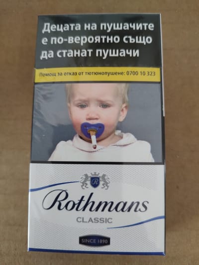 The image shows a pack of Rothmans Classic cigarettes. The front of the pack features a graphic warning about the dangers of smoking. In the foreground, a baby is depicted with a pacifier in its mouth, which is also holding a lit cigarette. The text on the pack is in Bulgarian and translates to "Children who smoke are more likely to become smokers." Below the image, the brand name "Rothmans" is prominently displayed, along with "CLASSIC" and "SINCE 1890." The packaging is predominantly white and blue with some gray elements. The background is a textured, brown surface, likely a table or floor. The scene is a close-up, top-down view of the cigarette pack. There is no indication of the location being Tervel, Bulgaria.