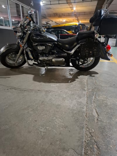 The image shows a dark grey and silver cruiser motorcycle parked on a concrete floor in an indoor parking garage. The motorcycle is positioned diagonally, facing towards the left side of the frame. It features chrome accents on the handlebars, headlights, engine, and wheels. Black leather saddlebags are attached to the rear of the motorcycle, and a black passenger backrest is mounted behind the seat. In the background, several yellow and black buses are visible, suggesting a public transportation hub or depot. The lighting in the garage is artificial, with ceiling lights illuminating the space. The concrete floor appears worn and has some cracks and wet patches. The overall scene suggests the motorcycle is parked and unattended in a utilitarian environment. There are no people visible in the image.
