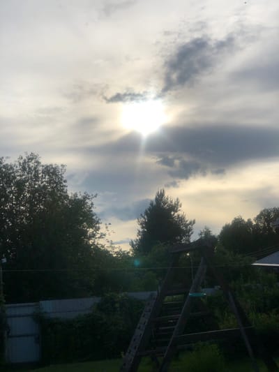 The image depicts an outdoor scene, likely a backyard, under a cloudy sky with the sun partially visible behind a thick cloud layer, casting rays of light downwards. In the foreground, a wooden swing set is prominently featured, showing signs of wear and tear, with a green circular object, possibly a swing seat or part of a toy, hanging from it. Beyond the swing set, dense greenery and trees form a natural backdrop. A light-colored fence runs along the left side of the frame, partially obscured by overgrown vegetation. In the distance, the silhouette of houses and more trees can be seen against the brighter sky. The overall impression is one of a rural or suburban setting, potentially in or near the city of Uriupino, Russia, during daylight hours, with overcast weather. There are no people or animals visible in the image.
