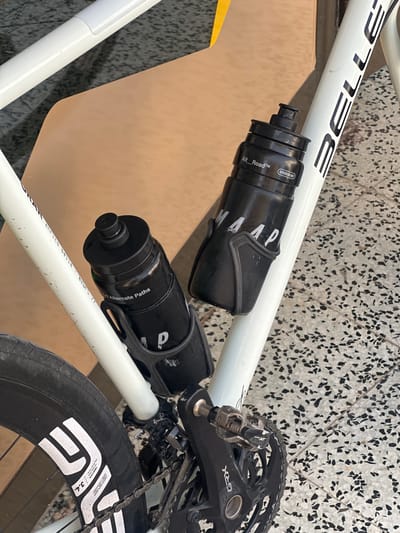 The image shows a close-up view of two black water bottles mounted on the white frame of a bicycle. The bicycle frame has black lettering that reads "BELLE" on it. One water bottle, closer to the viewer, is inscribed with "MAAP" and the other, slightly further back, has "Alt_Road™" and "OUTDOOR USE ONLY" printed on it. The bicycle is positioned on a patterned tiled floor. A portion of the rear wheel with "SES 3.4" branding is visible. The scene appears to be outdoors, possibly in a urban setting given the tiled ground. The overall impression is one of a detail shot of a bicycle's accessories, with no people or specific activity depicted. The location is La Llacuna, Spain.