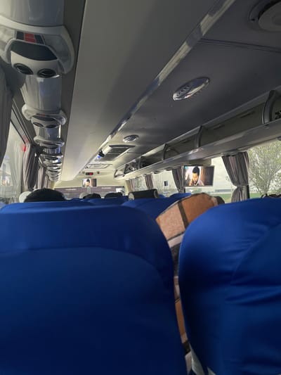 The image depicts the interior of a Yutong passenger bus, likely traveling along a route in Karmana, Uzbekistan, based on the displayed name. The bus features blue upholstered seating and individual entertainment screens mounted above each row. The lighting fixtures and overhead storage compartments are visible. Some passengers are seated, with hats and blankets. The exterior is seen through the windows, where trees are visible.