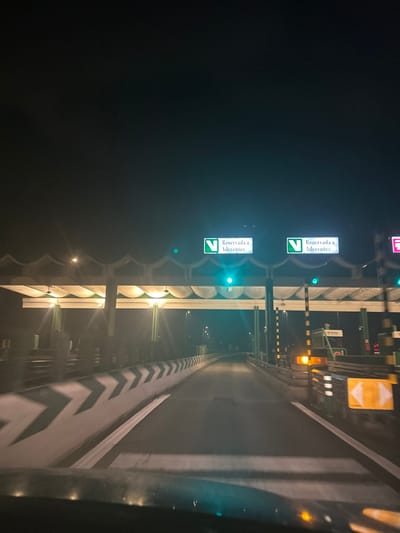 Nighttime highway tollbooth, illuminated with green lights and overhead signs that read "Reservada a Aderentes." An empty toll lane stretches out, defined by chevron-patterned barriers. Beyond the green lights, other tollbooths show orange and yellow lights. The scene captures the stillness of the highway under the dark sky.