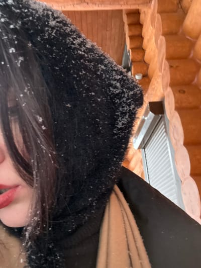 The image presents a close-up, rotated view, focusing on the lower portion of a person's face and their winter attire, set against a backdrop of wooden structures. The orientation of the image is tilted roughly 90 degrees counter-clockwise from a standard portrait view.
The central subject is a person whose dark hair and part of their face are visible on the right and lower right side of the frame. Their dark hair, possibly long, is partially covered by a black, seemingly furry or plush hood or hat. Small, white flakes, characteristic of snow or ice, are scattered across the black material of the hood and on some strands of the person's dark hair. The lower part of the person's face is visible, showing their chin, a hint of a cheek, and distinct red lips that are slightly parted. To the far right, a section of beige or light brown fabric, possibly a scarf or part of a jacket collar, is visible, tucked underneath or alongside the black hood.
The background consists of architectural elements, suggesting an outdoor or semi-sheltered environment. Above the person's head (which would be to their left if the image were upright), there is a structure made of light-colored, possibly natural wood logs. These logs are horizontally stacked, showing their rounded profiles, consistent with log cabin construction. A small, white, circular object, potentially a light fixture or sensor, can be faintly seen attached to one of the wooden logs in the upper-left area. Between the log structure and the person's headwear, a light grey or white corrugated or slatted surface is visible, likely forming an overhang or ceiling. Further back on the left side, another wooden surface, possibly planks, is visible.
The presence of snow flakes on the person's attire and hair strongly indicates a cold, snowy, outdoor winter setting. The lighting appears bright and natural, suggesting daytime. No discernible text is visible in the image.