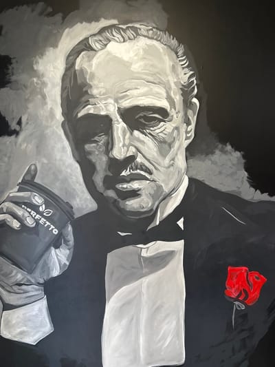 The image is a painting of Marlon Brando as Vito Corleone from The Godfather. He is depicted in a grayscale portrait, wearing a tuxedo and holding a cup with the "PERFETTO" logo. A single red rose is pinned to his lapel. The background is dark and abstract, with some wisps of gray paint suggesting smoke or shadows. The painting is rendered in a loose, expressive style. There is no indication in the image that this painting is located in Yakutsk, Russia.